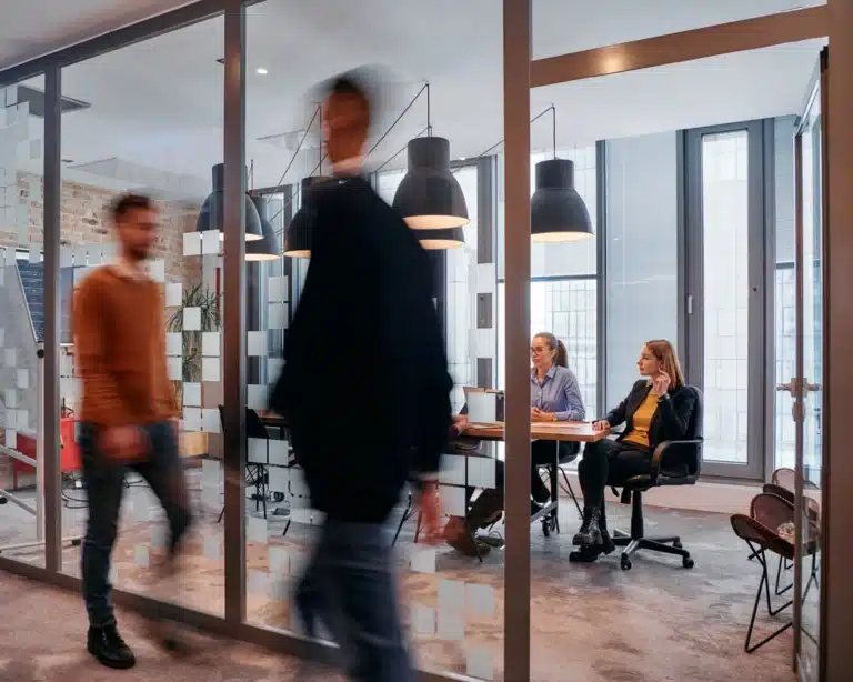 A group of young business professionals walk down the corridor next to their office, while their colleagues collaborate inside.