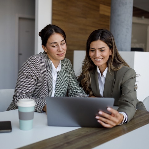Two young businesswomen are smiling and working together on a digital tablet in a modern office.